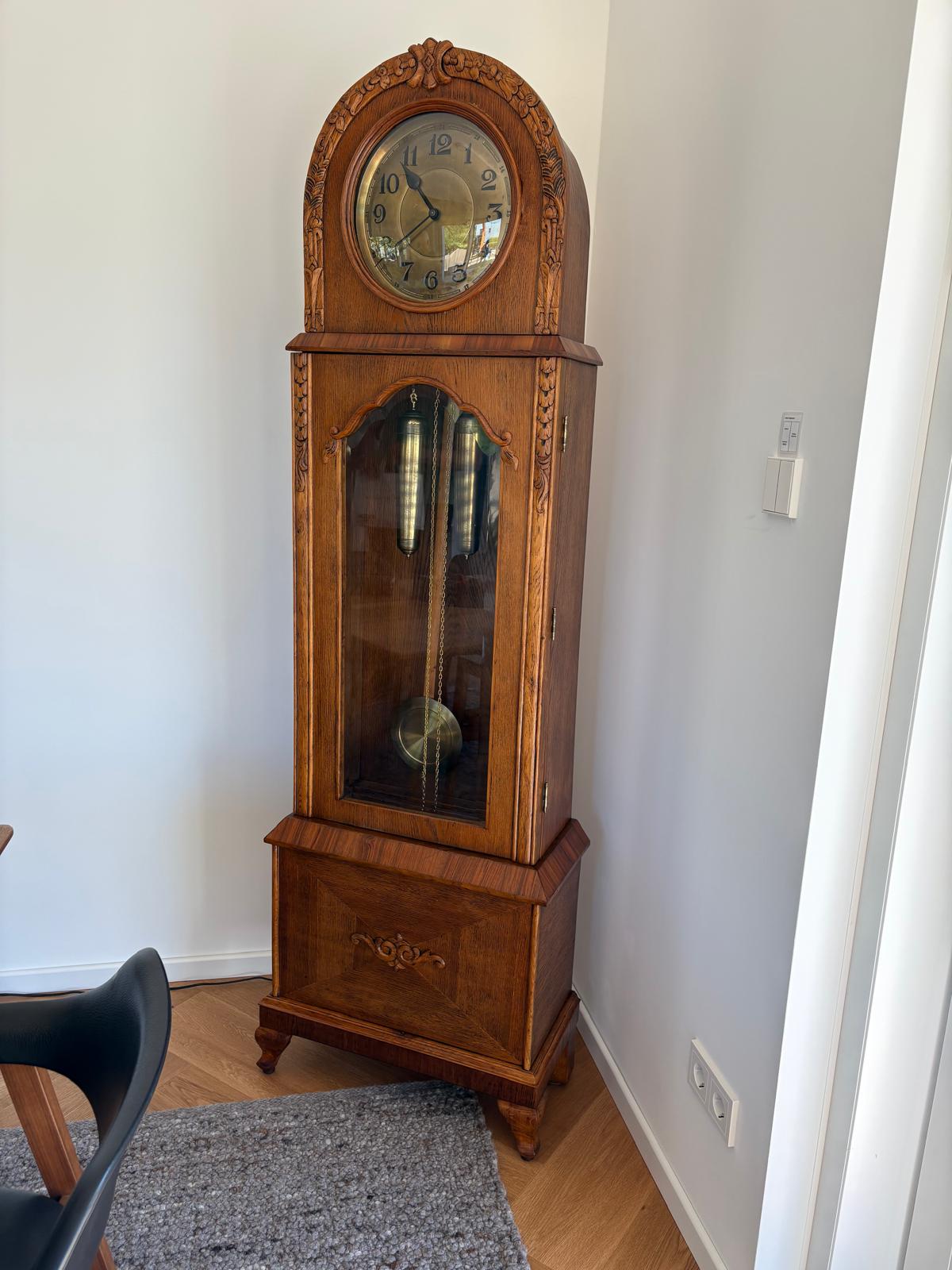 Fertig restaurierte antike Standuhr in heller Holzoptik, aufrecht in einem Wohnraum und vollständig von vorn zu sehen.
