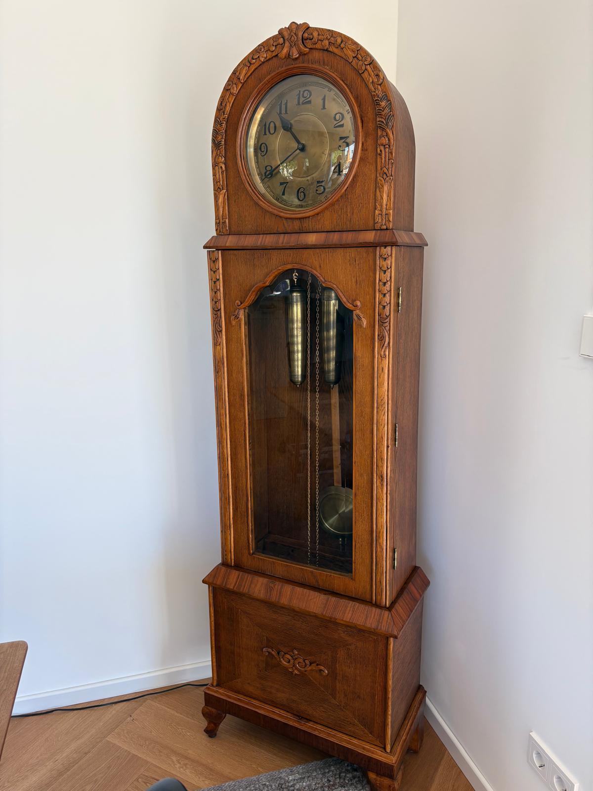 Vollständig restaurierte antike Standuhr aufrecht im Wohnraum, frontal mit Ziffernblatt, Pendelbereich und Sockel fotografiert.